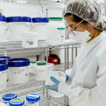 A person in full protective gear reading a white container with a metal shelf of other containers behind them.