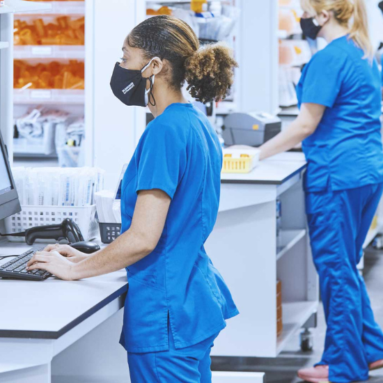 Woman in blue scrubs and a black mask standing and working at a computer.