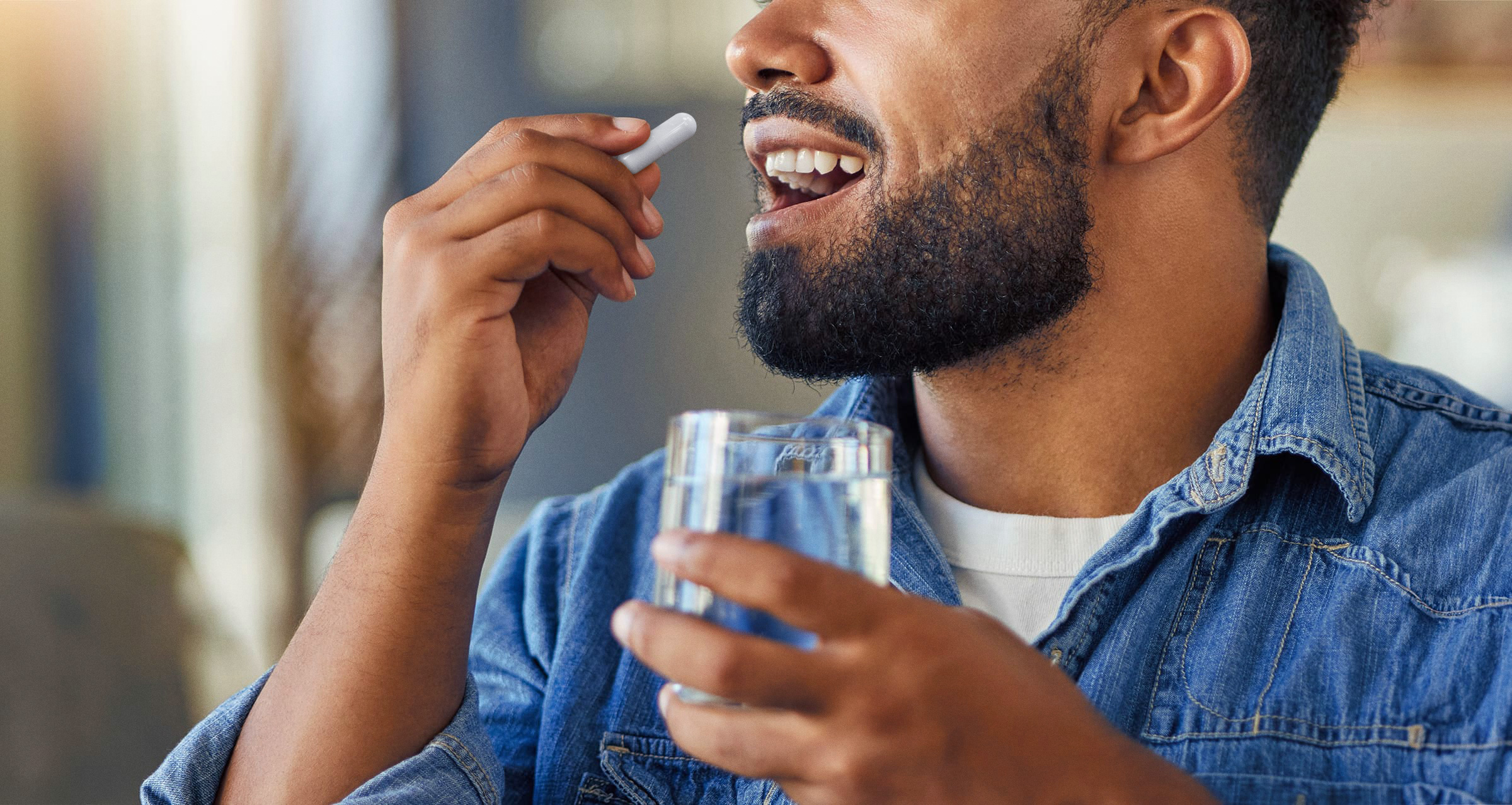 A man with a black beard in a denim shirt taking a white pill while holding a glass of clear liuid.