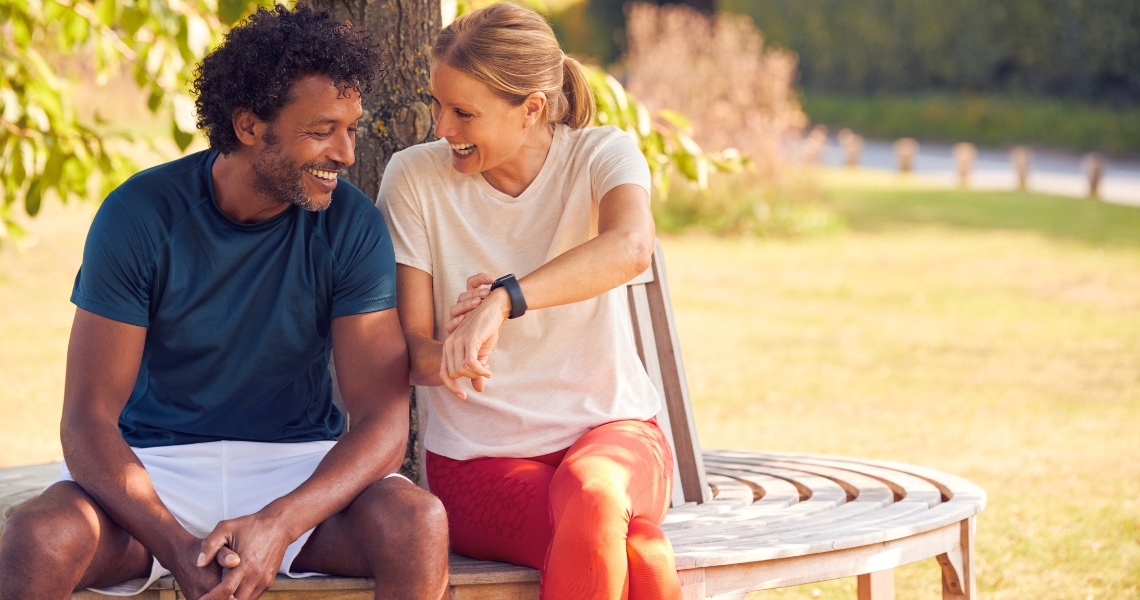 A woman with blonde hair showing her smart watch to an African American man while both sitting on a bench outside.