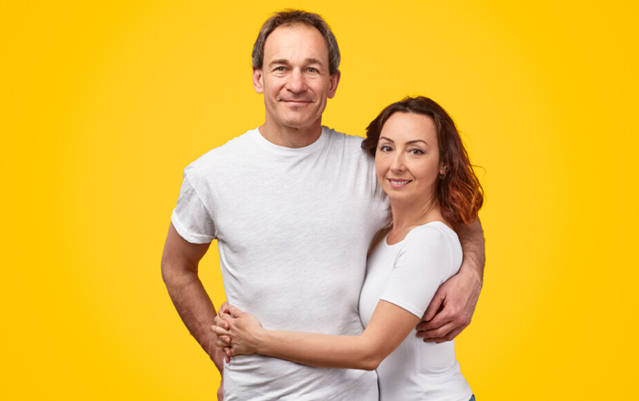 Yellow background. Middle aged man and middle aged woman in white t-shirts with arms around each other facing forward.