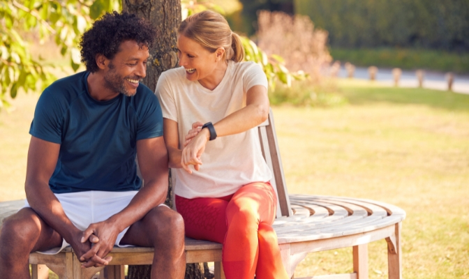 A woman with blonde hair showing her smart watch to an African American man while both sitting on a bench outside.