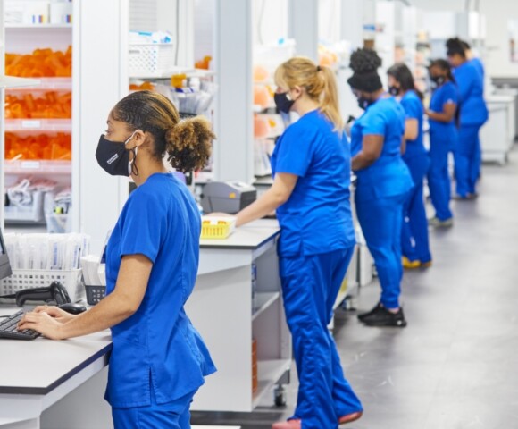 A row of technicians in blue scrubs working at individual work stations.