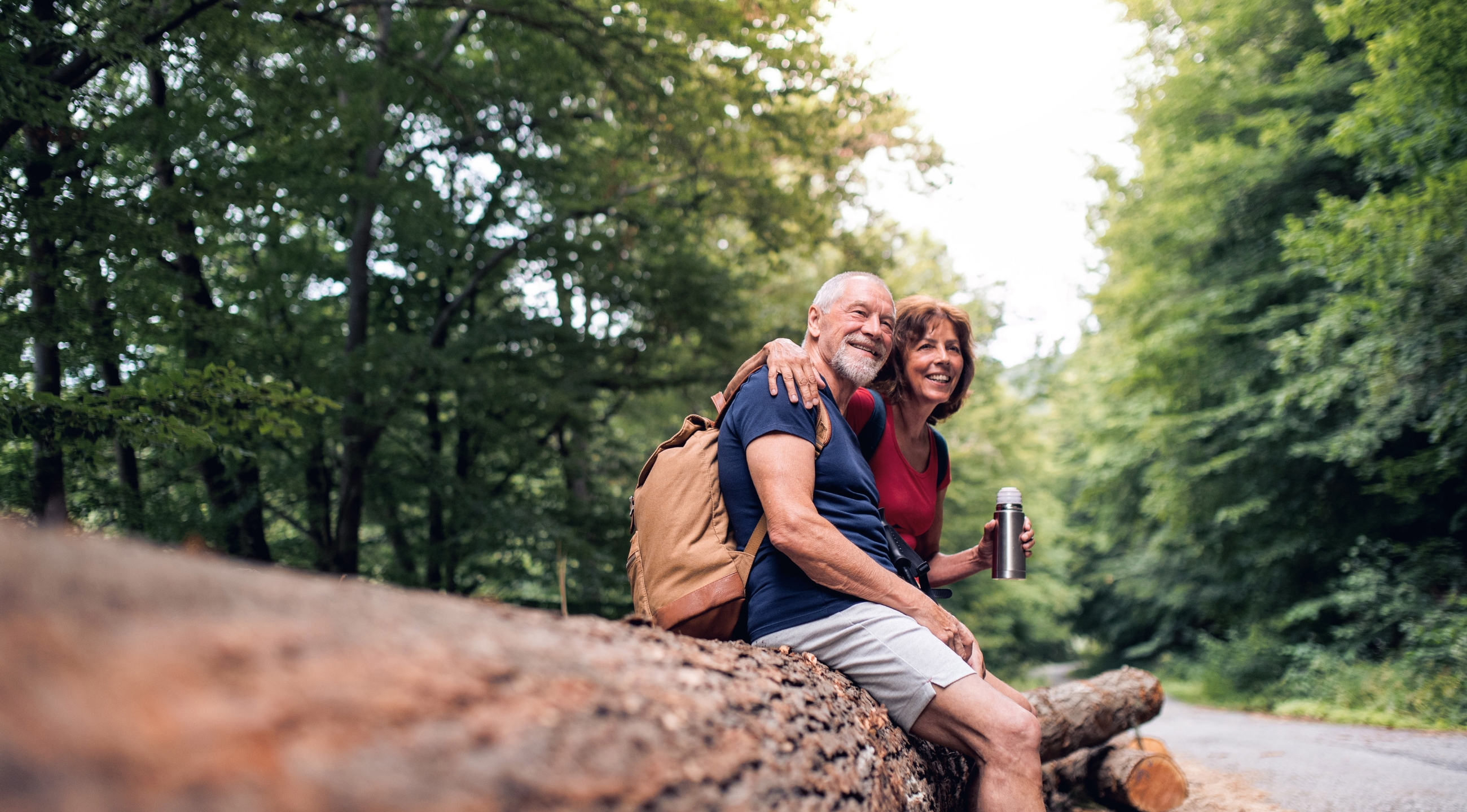 An older aged man and woman sitting arm and arm outdoors on a fallen tree.