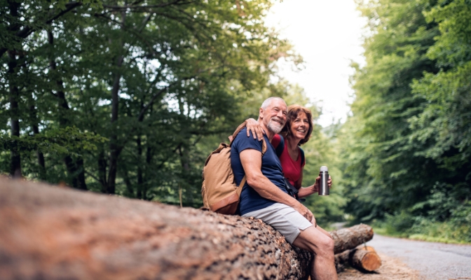 An older aged man and woman sitting arm and arm outdoors on a fallen tree.