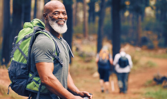An African American man with a gray hair wearing a green backpack and gray shirt hiking in the woods on a trail.