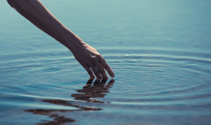 A hand tracing its fingers over the top of a blue body of water causing ripples.