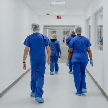 A view from the back of three people in navy blue scrubs and protective gear walking down a white hallway.