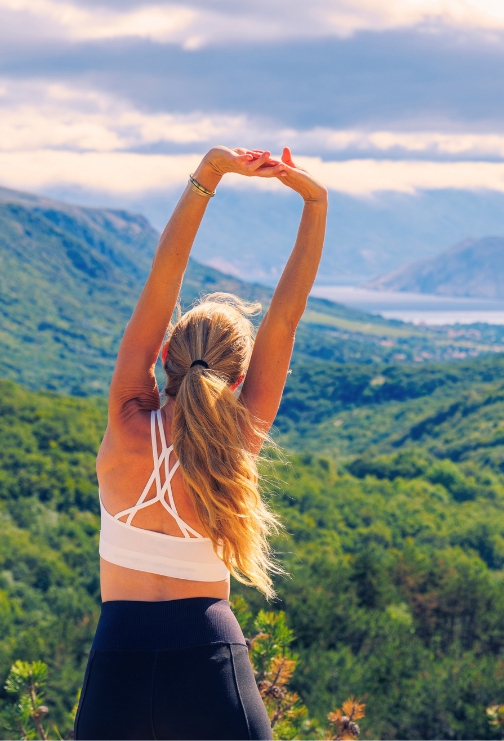 A woman in a white workout top and black leggings stretching with mountains in front of her.