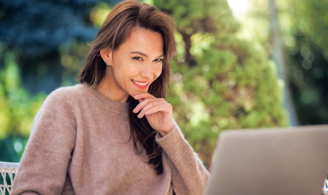 A middle-aged woman with brown hair using her laptop outdoors.