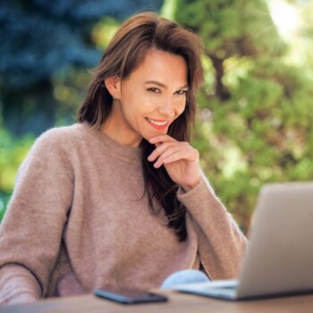 A middle-aged woman with brown hair using her laptop outdoors.