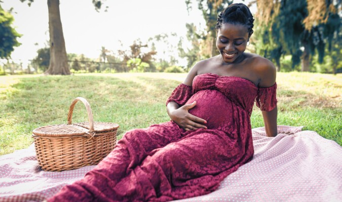 A pregnant woman in a maroon dress laying down outside holding her stomach.