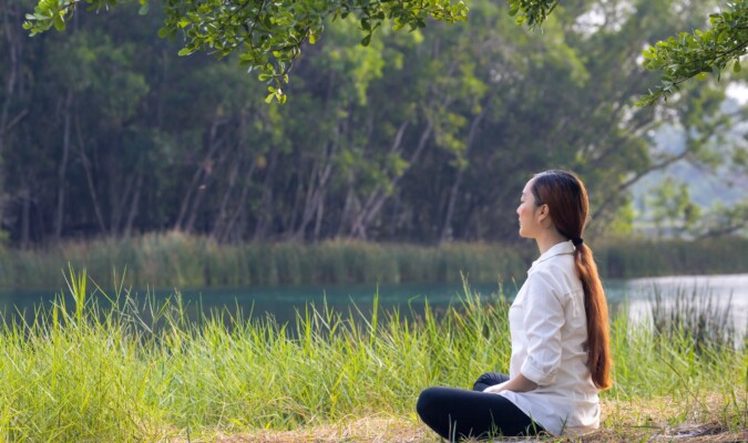 A woman meditating outdoors by a body of water.