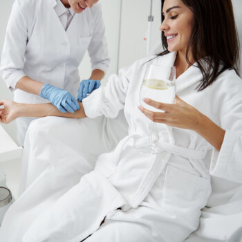 Woman in robe with glass of water in her hand receiving an IV from a nurse in blue disposable gloves.