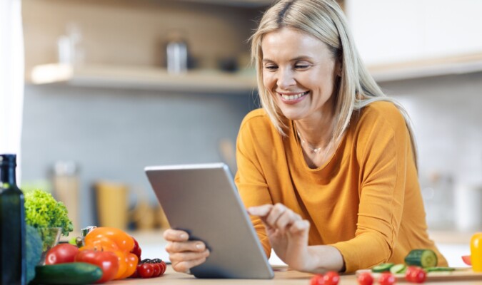 A woman using a tablet in the kitchen surrounded by produce.