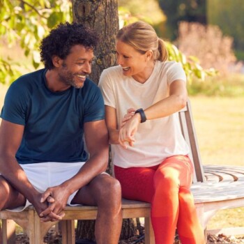 A woman with blonde hair showing her smart watch to an African American man while both sitting on a bench outside.