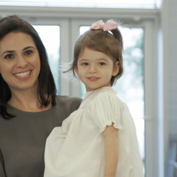 A smiling woman holds a young girl with a pink bow in her hair. They are standing in front of a window with natural light coming in. The girl is wearing a white dress and looking at the camera, while the woman stands behind her, smiling warmly.